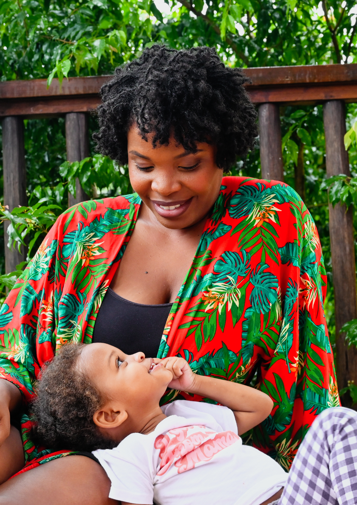 Une femme souriante portant une robe colorée avec un motif tropical, assise à l'extérieur, interagissant joyeusement avec un jeune enfant qui sourit et joue.